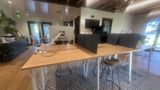 Long shared work tables with monitors and low privacy screens, under the exposed wood ceiling beams, with daylight from the street-side windows